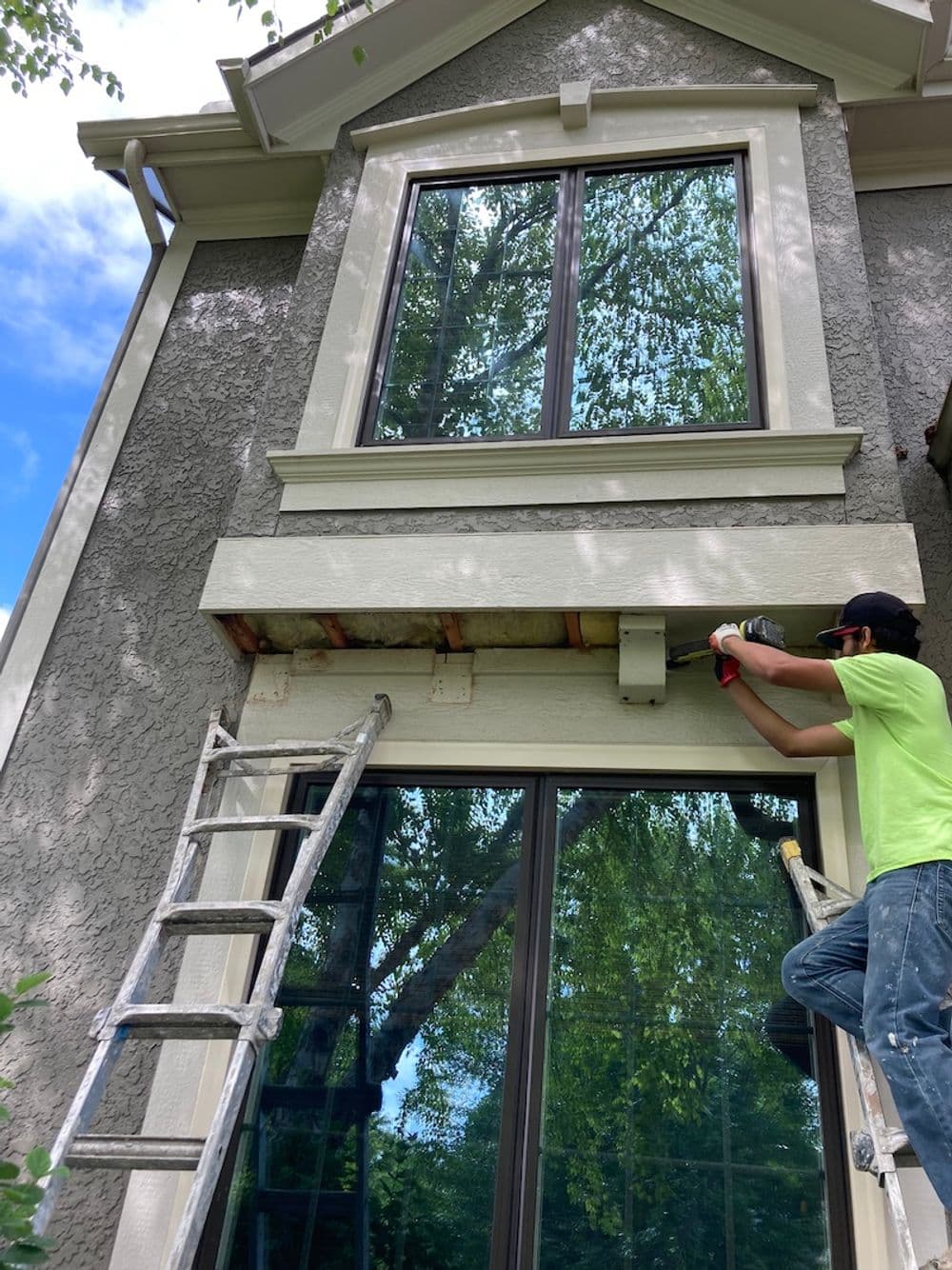 Worker using a power tool on a home's exterior above a ladder, with large windows and trees in view.