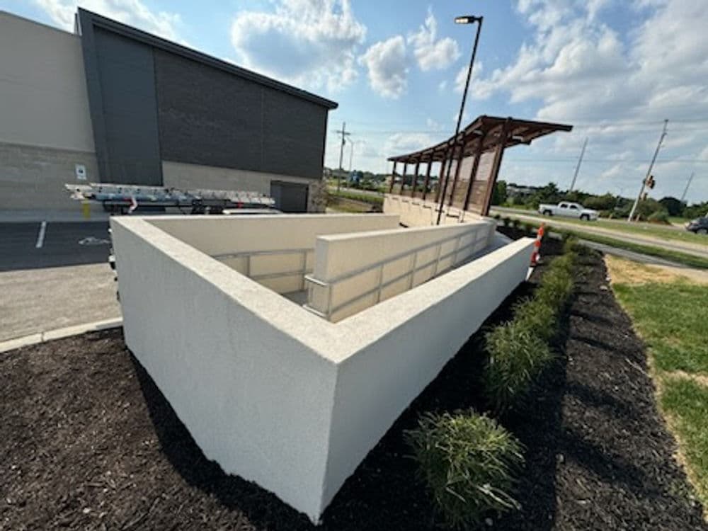 Modern concrete structure with landscaping, parking lot, and wooden shelter in background.